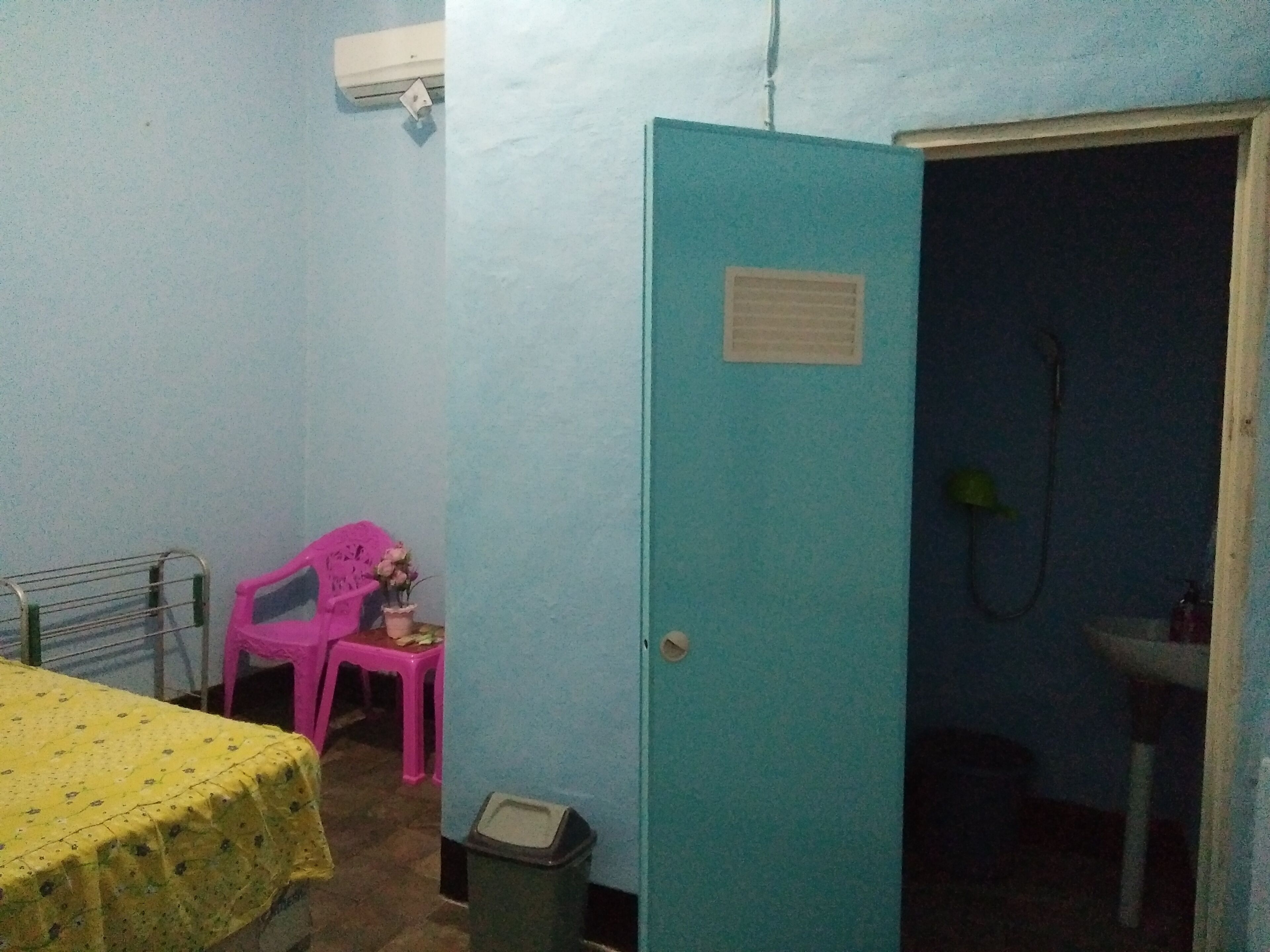 A small room with a bed, a pink chair, a trash bin, and a door leading to a bathroom.
