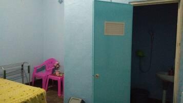 A small room with a bed, a pink chair, a trash bin, and a door leading to a bathroom.