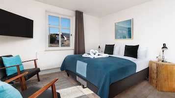 A modern hotel room with a large bed, a flat-screen TV mounted on the wall, a window with curtains, a small wooden side table, and a view of the outside.