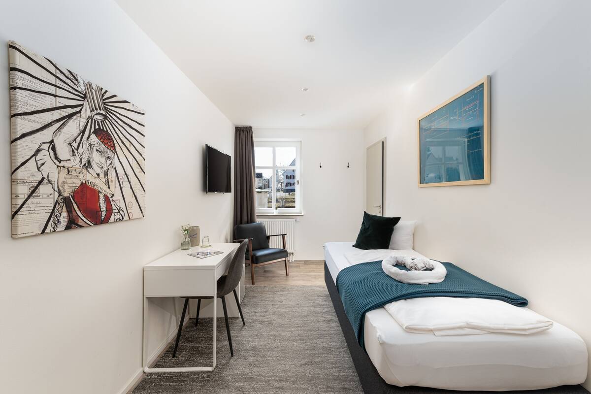 A compact hotel room with a single bed, a desk, and a chair. A large abstract painting adorns the wall, and a window offers a view of the city.
