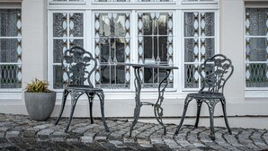 Een wit gebouw met een groot rondboogvenster met glas-in-lood. Twee sierlijke metalen stoelen en een kleine tafel staan ervoor neergezet.