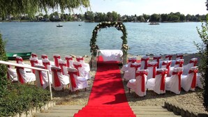 A wedding ceremony setup with a red carpet leading to chairs with white covers and red sashes, a floral arch, and a lake view. - Hotel Zátoka (Senec)