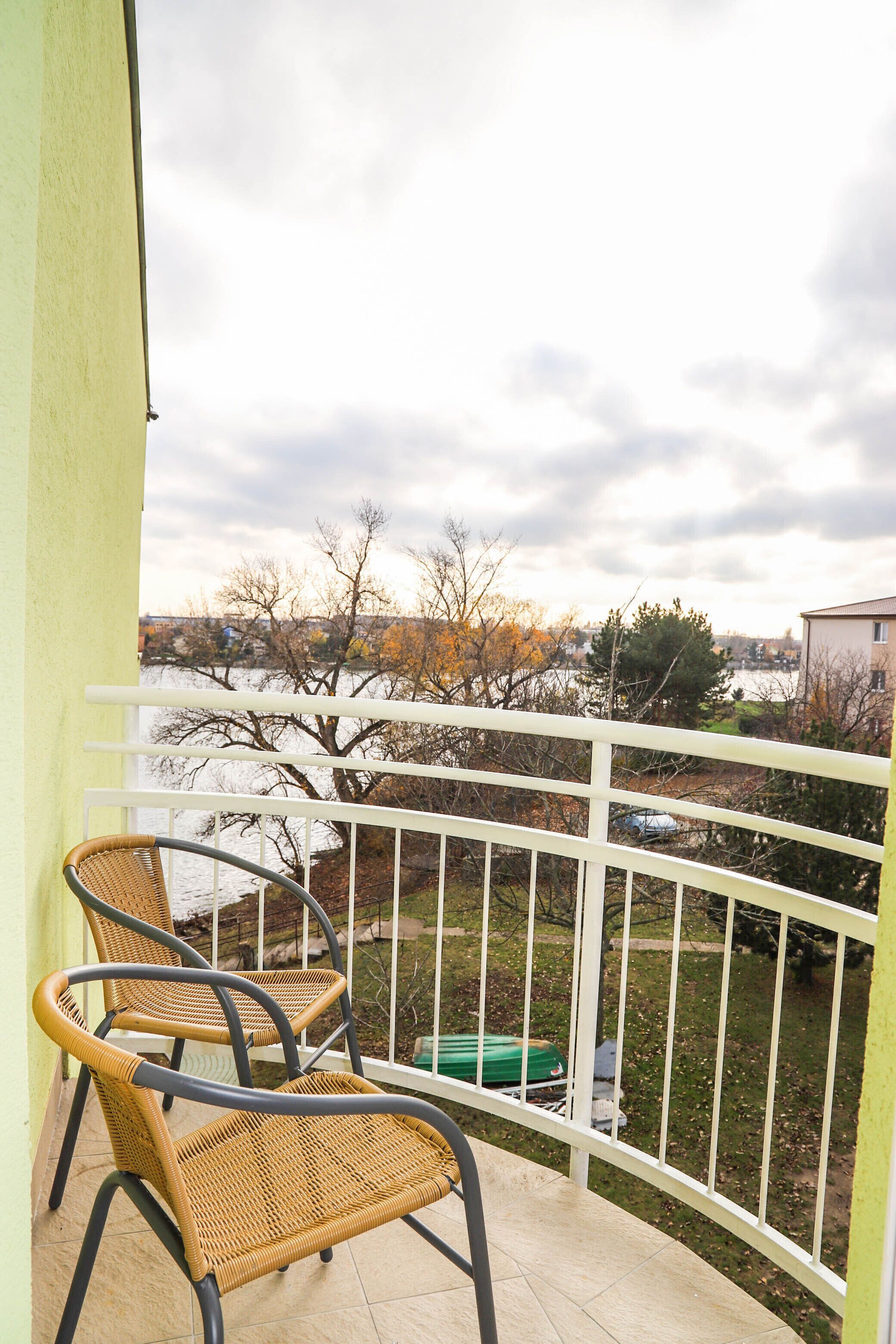Un balcon avec deux fauteuils en osier, une balustrade blanche et une vue sur des arbres et une étendue d’eau.