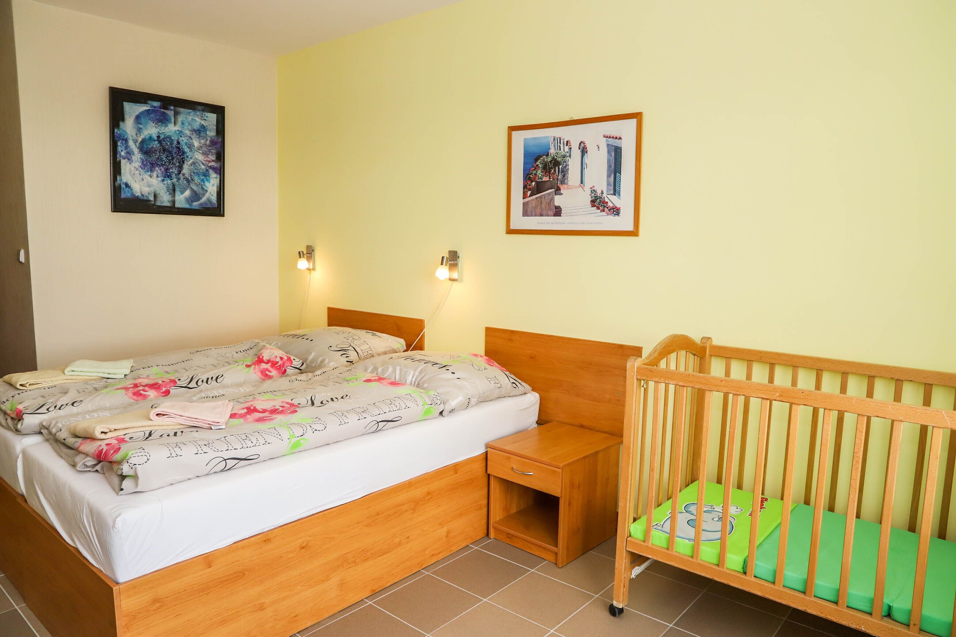 A bedroom with a bed, a nightstand, a crib, two wall-mounted lights, and two framed pictures.