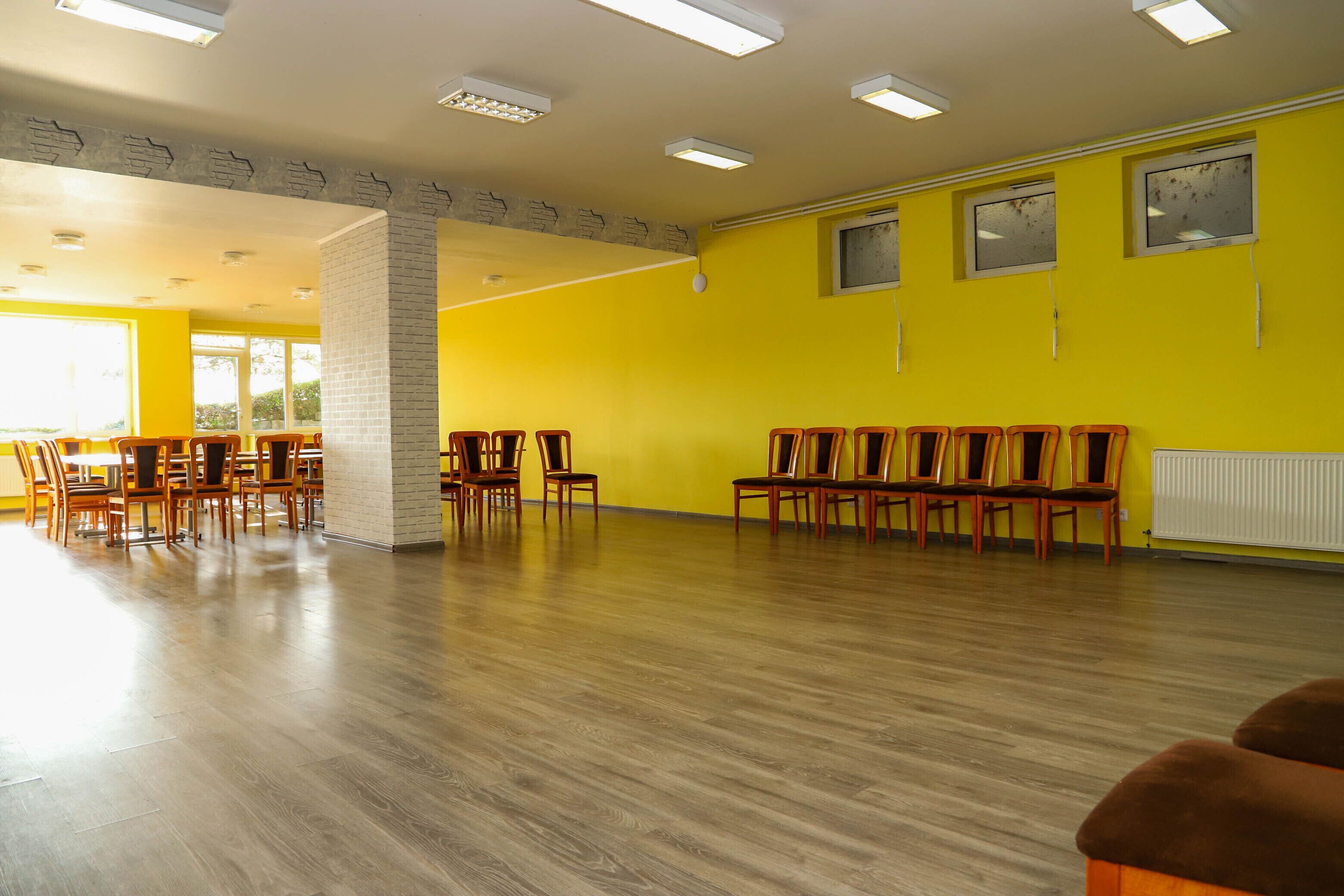 A spacious room with wooden flooring, yellow walls, and multiple wooden chairs arranged for seating.