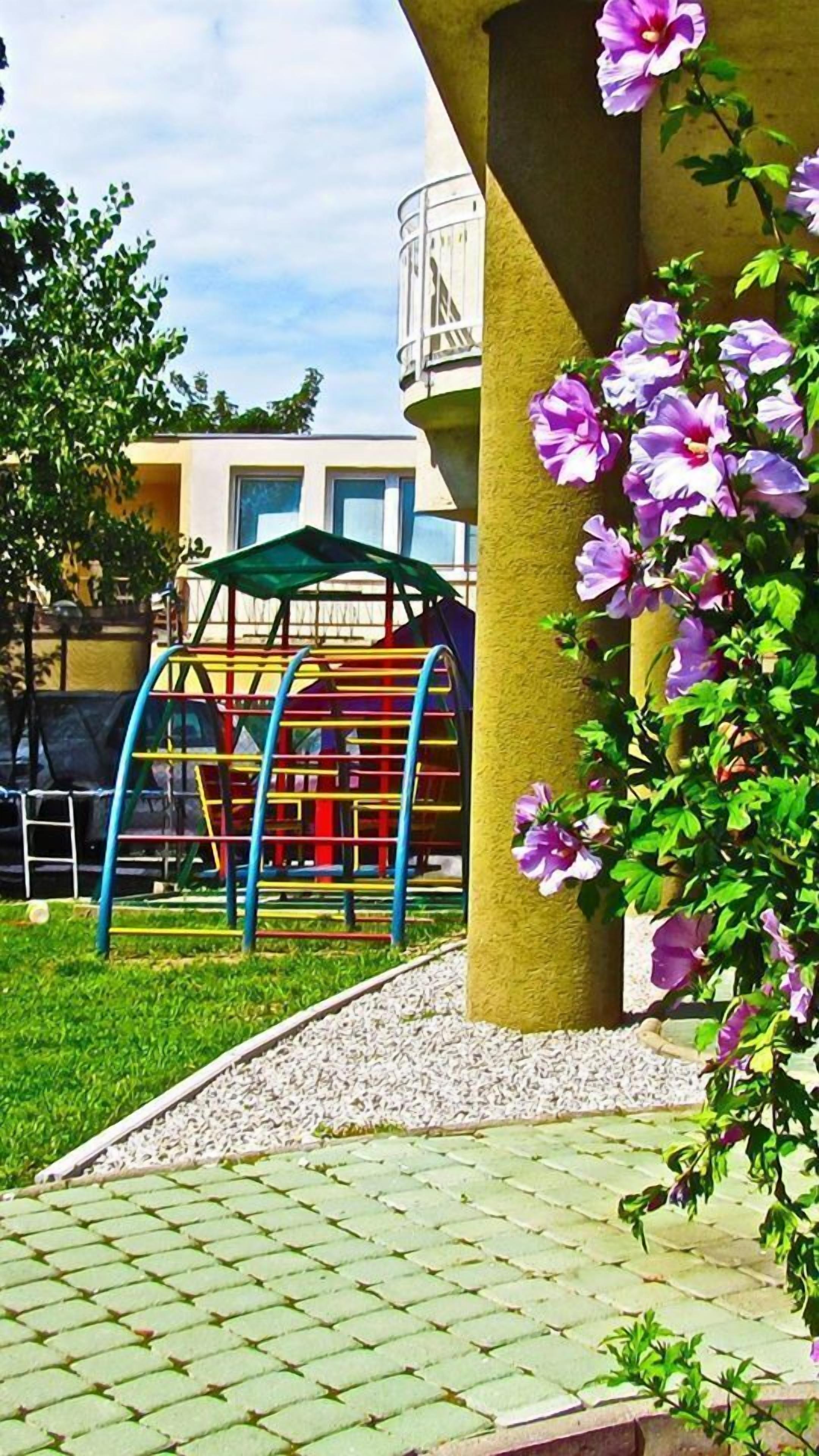 Un parque infantil con una estructura para escalar y una zona de descanso cubierta, rodeado de una superficie pavimentada y plantas con flores.