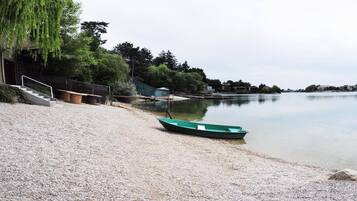 Una playa de grava con un bote verde, una escalera que baja hacia el agua y una fila de árboles y edificios al fondo.