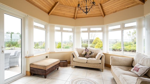 A sunlit room with a high ceiling, large windows, a sofa, a bench, and a patterned rug. - Drum Manor (Hillsborough)