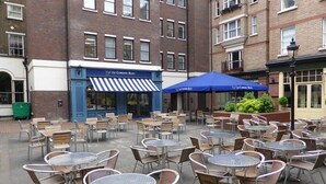 Een terras met tafels en stoelen, een blauw zonnescherm en een gebouw met een gestreept zonnescherm.