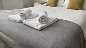 Een net opgemaakt bed met witte en gouden kussens, een grijze hoofdbord, en twee opgerolde witte handdoeken op het bed.