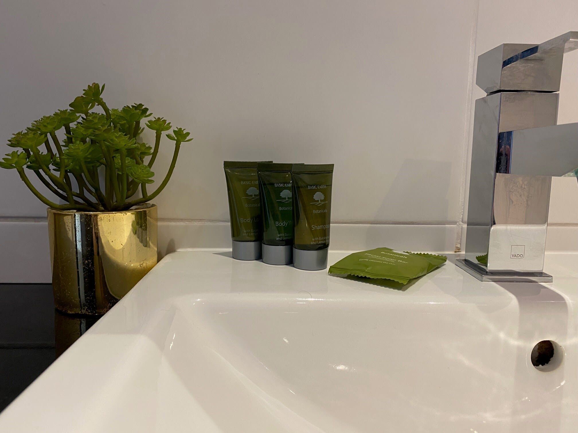 A bathroom sink with a gold vase containing green plants, three green bottles of body lotion, and a green packet of botanical soap.