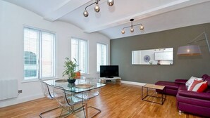 Un salon moderne avec une table à manger en verre, des chaises transparentes, un canapé violet, un téléviseur et une grande fenêtre équipée de stores.