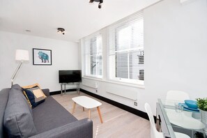 Ein modernes Wohnzimmer mit einem grauen Sofa, einem kleinen weißen Tisch, einem Glastisch mit Stühlen, einem Flachbildfernseher und einem großen Fenster mit Jalousien.