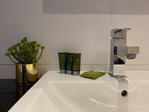 A bathroom sink with a modern faucet, a gold vase with green plants, and three green bottles of body wash. - 402H Warehouse Conversion 1 bed plus sofa bed (London)