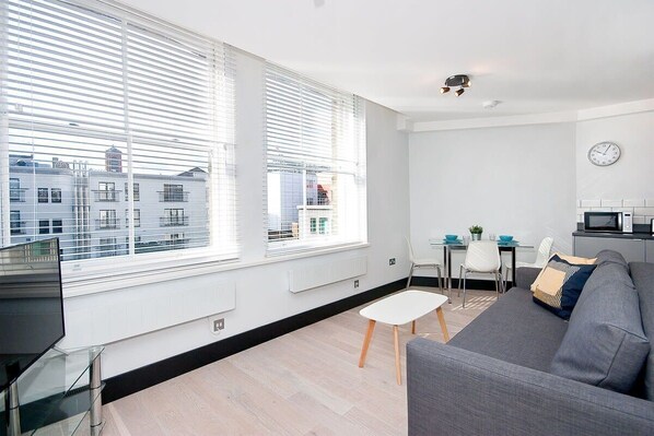 A modern living room with a grey sofa, a small white table, and a dining area with a white chair. Large windows provide a view of buildings outside. - 402H Warehouse Conversion 1 bed plus sofa bed (London)