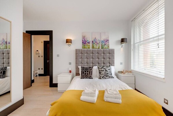 A hotel room with a large bed, a headboard with a tufted design, two bedside tables, a nightstand, a mirror, and a window with blinds.