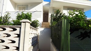 Un étroit sentier mène à un bâtiment blanc, bordé d’une clôture verte et agrémenté de plantes en pot.