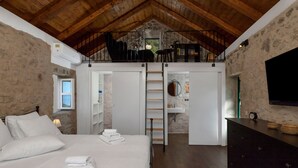 Un dormitorio con techo de madera, paredes de piedra, una cama con ropa blanca, un armario y un televisor montado en la pared.