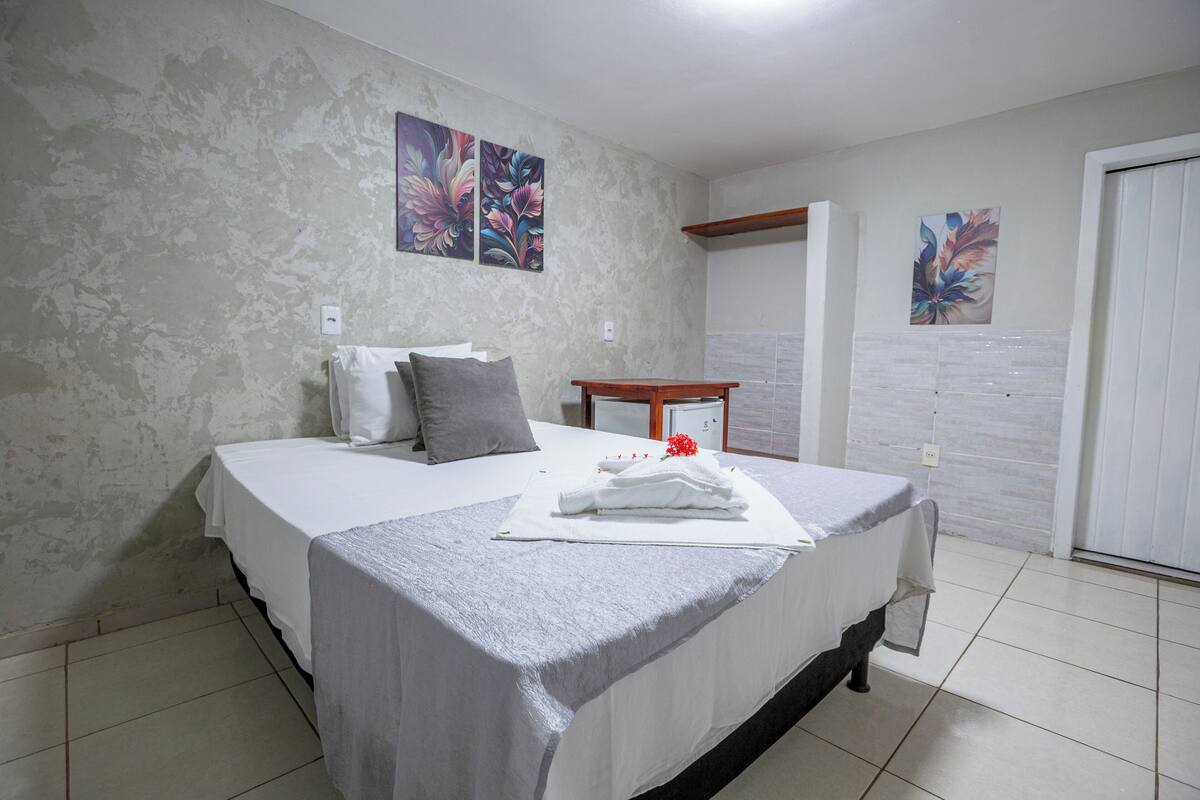 A neatly made bed with a white bedspread and pillows, a small wooden table with a white cloth, and two floral paintings on the wall.
