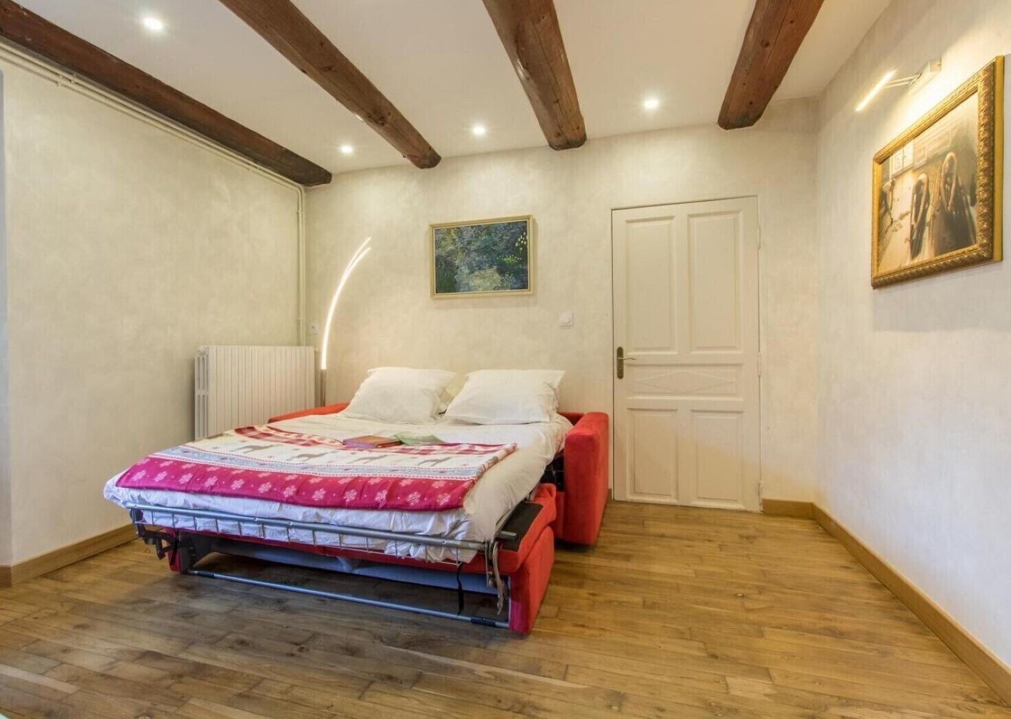 A bedroom with a bed, a radiator, a door, and a painting on the wall.