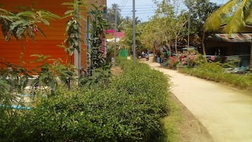 Un camino rural con un edificio rojo a la izquierda, rodeado de vegetación y algunas casas al fondo.