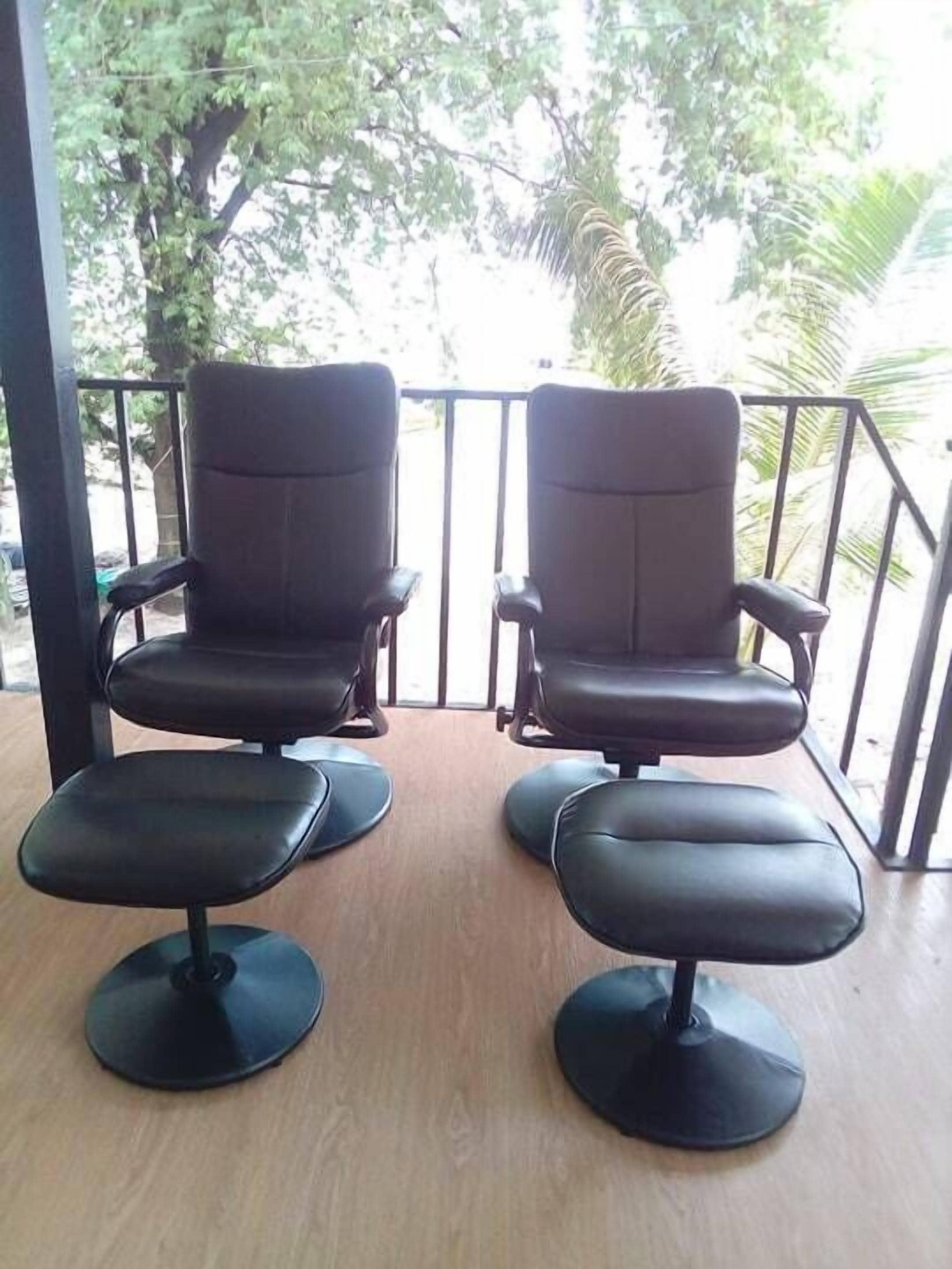 Dos sillones de cuero negro con reposapiés en una terraza de madera, con vista a un jardín con árboles y una barandilla.