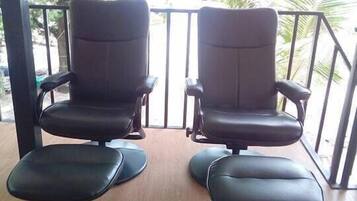Dos sillones de cuero negro con reposapiés en una terraza de madera, con vista a un jardín con árboles y una barandilla.