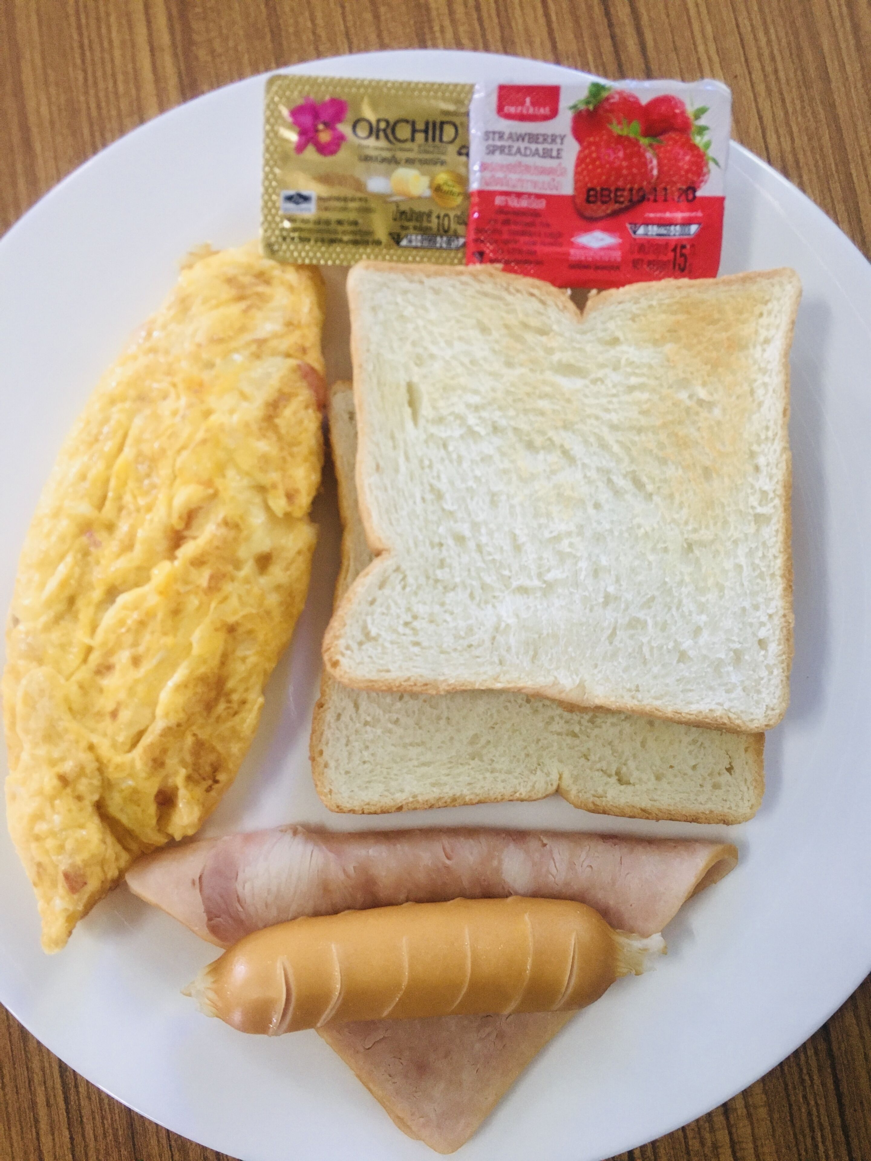 A plate with an omelette, two slices of white bread, a slice of ham, and a packet of strawberry spreadable.