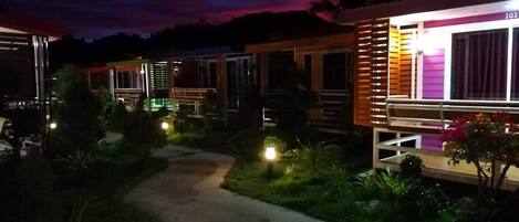 A row of modern, brightly lit cabins with a pathway leading through a landscaped garden.