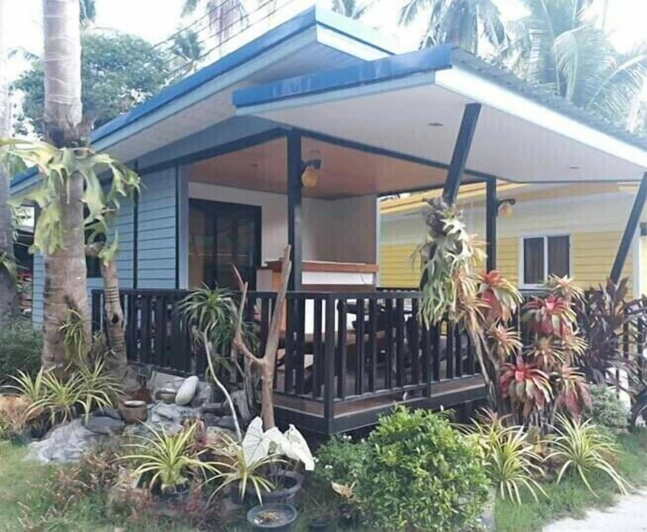 A two-story house with a balcony, surrounded by tropical plants and a small garden.