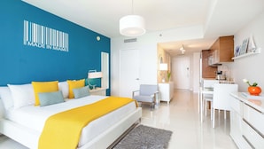 A modern hotel room with a large bed, a bright blue accent wall, a kitchen area, and a dining space.