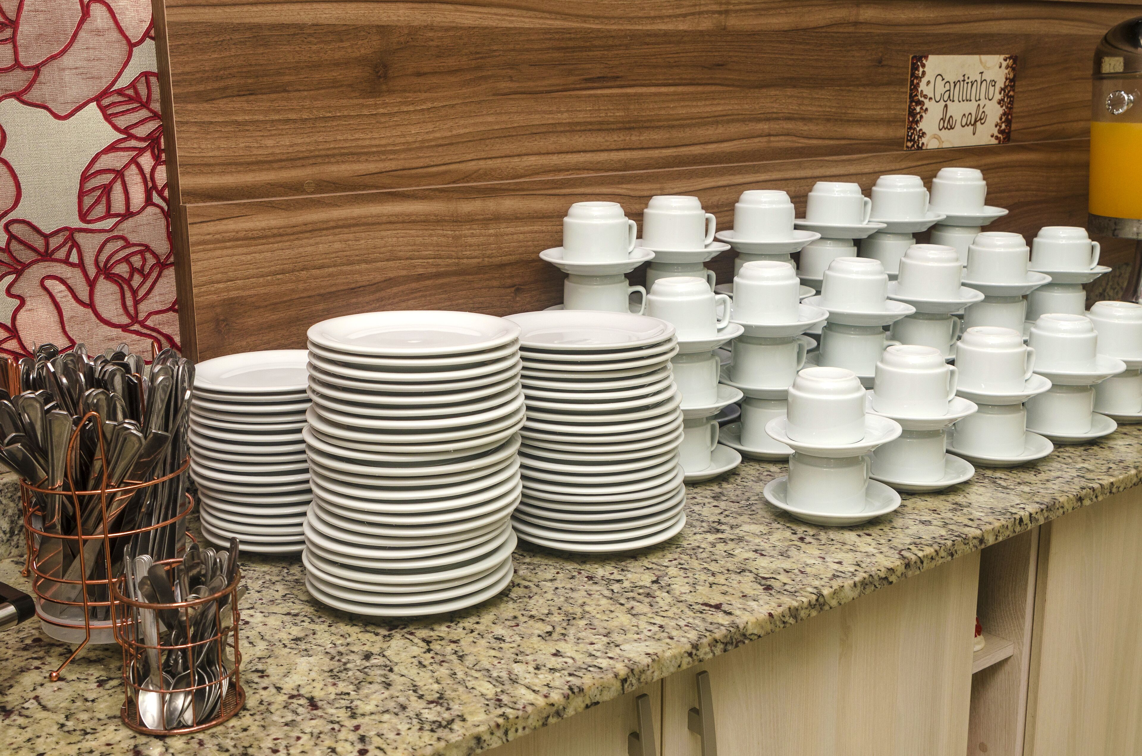 Pilas de platos y tazas blancos sobre una encimera de granito, con una cafetera y un letrero que dice "Cantinho do Café" al fondo.