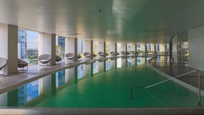 Ein Innenpool mit klaren Glaswänden, Korbstühlen und Blick auf die Stadtlandschaft.