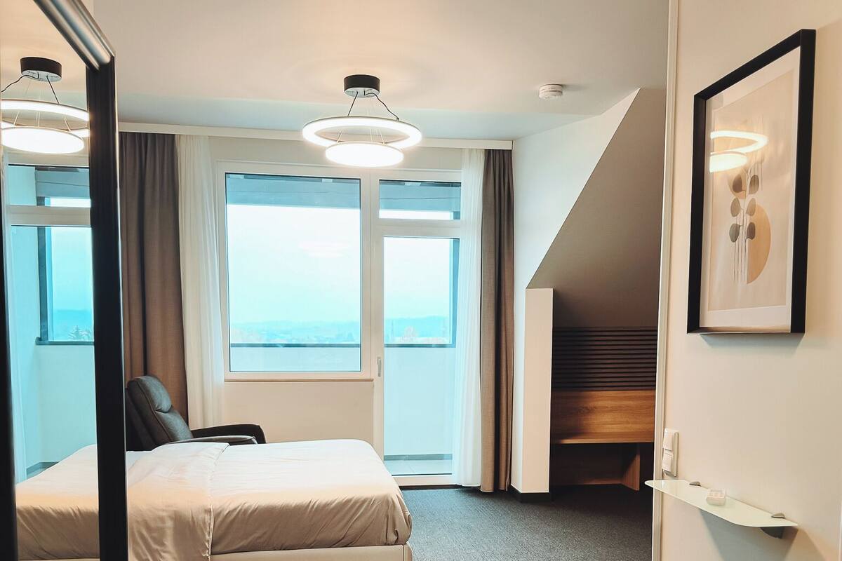 A modern hotel room with a large bed, a chair, a desk, a desk lamp, a framed picture, and a view of the city through the window.