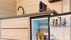 A compact kitchen with a built-in refrigerator, wooden cabinetry, and a small shelf with snacks and drinks. - F Hotel 24h Check-In (Hoersching)