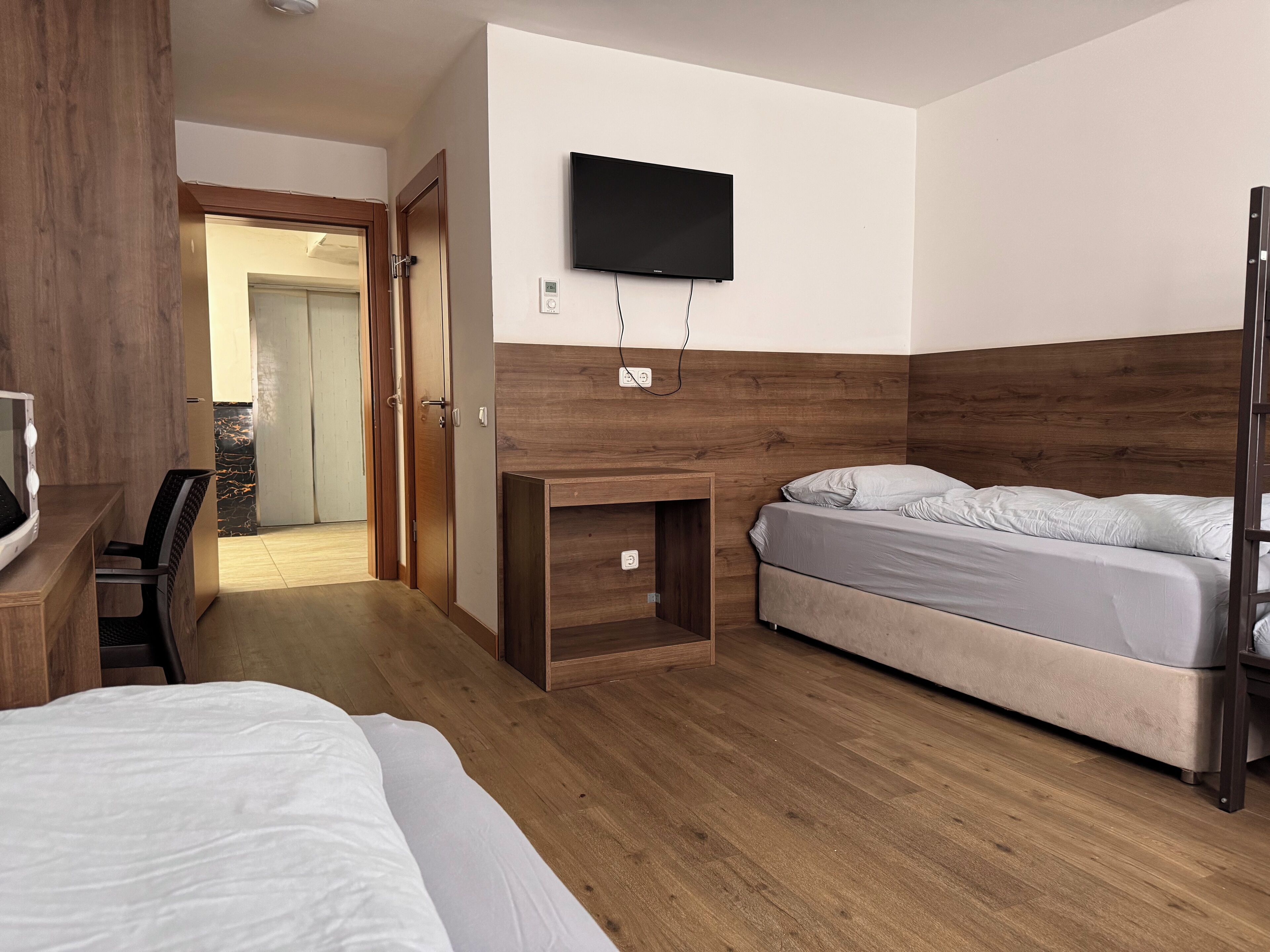 Une chambre d’hôtel avec un plancher en bois, un lit, un bureau avec une chaise, un téléviseur fixé au mur et une porte donnant sur une autre pièce.