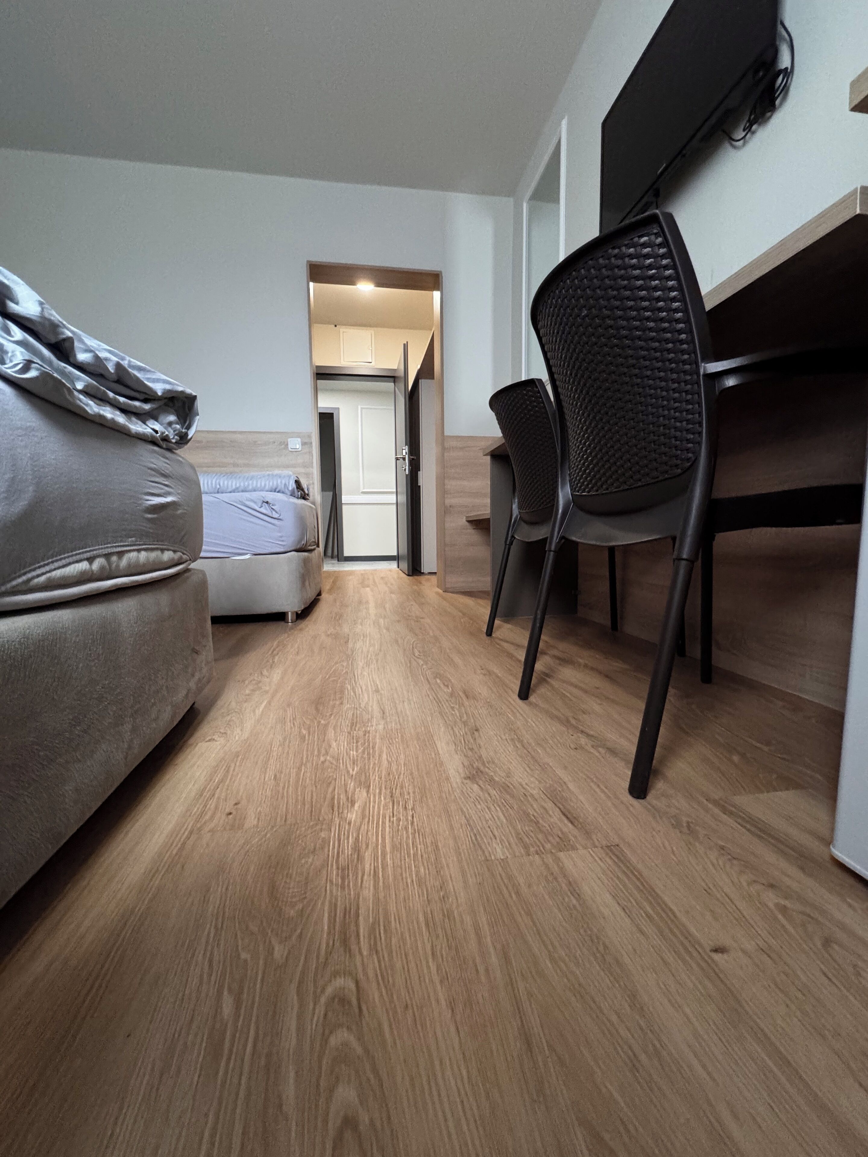 A modern, compact room with a wooden floor, a bed, a desk with a chair, and a door leading to another room.