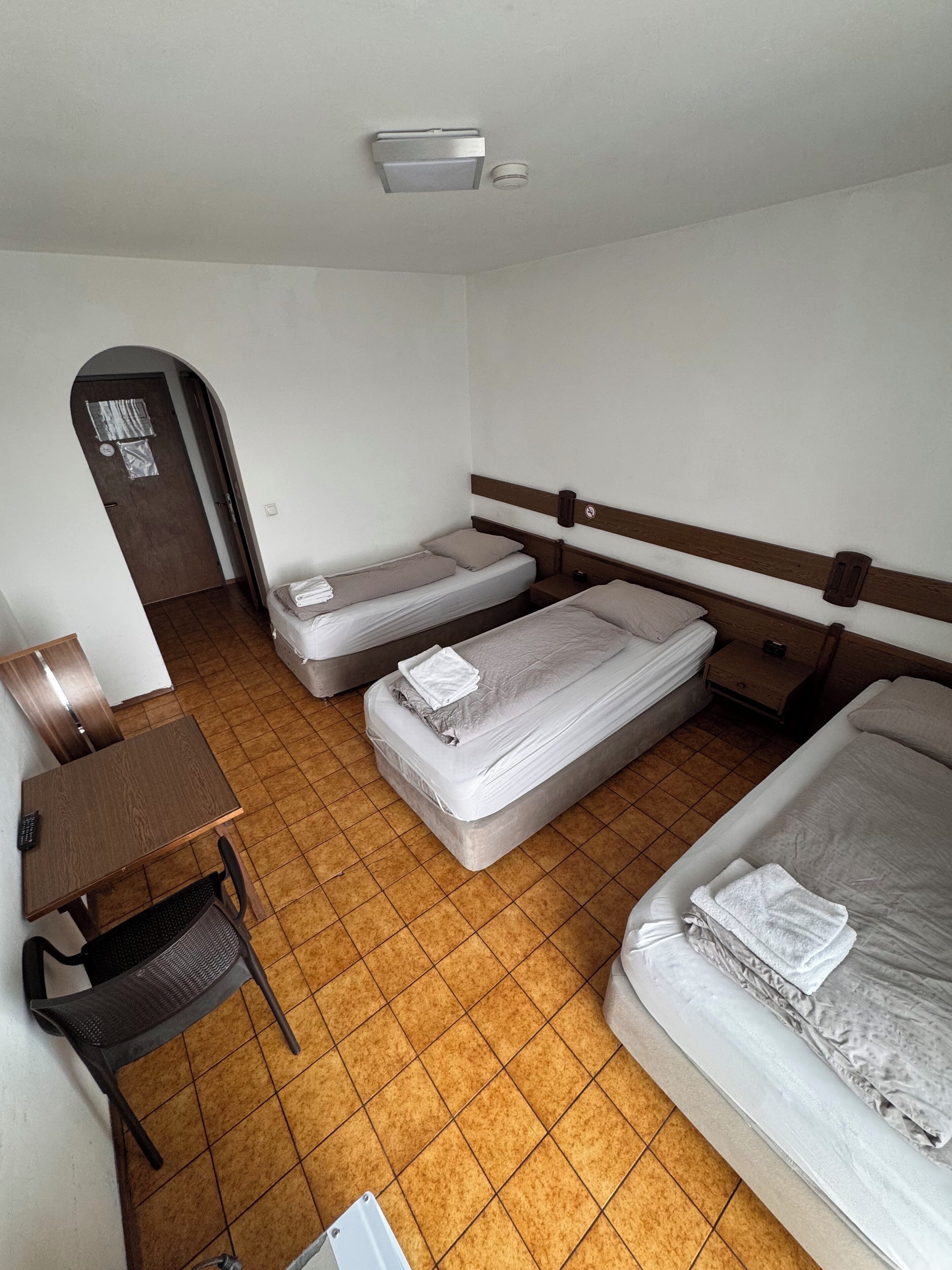 A hotel room with two single beds, a larger bed, a desk with a chair, and a small table.