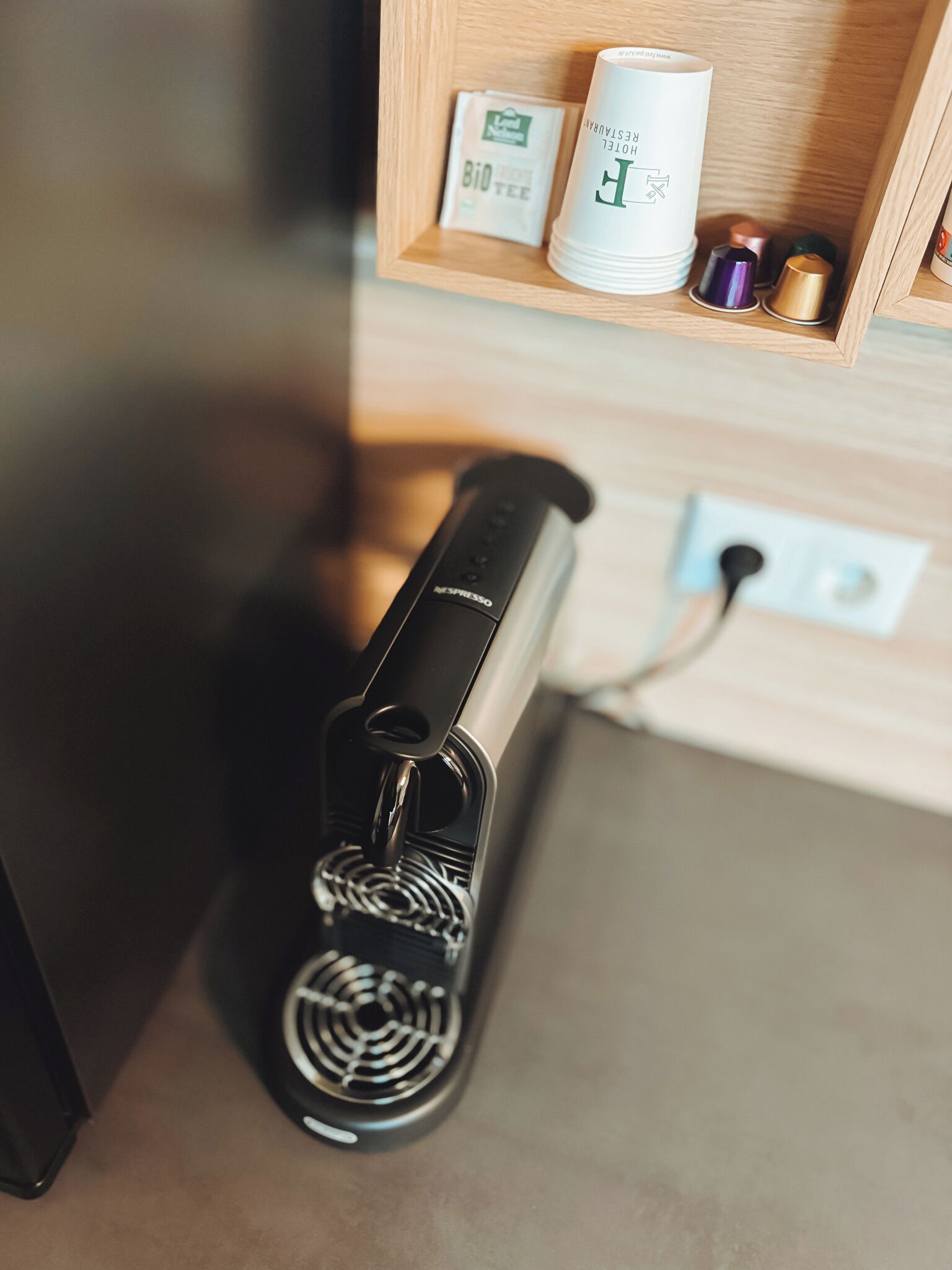A coffee machine with a power cord plugged in, a wooden shelf with a coffee cup, a tea bag, and a small box labeled "Bio Tea".