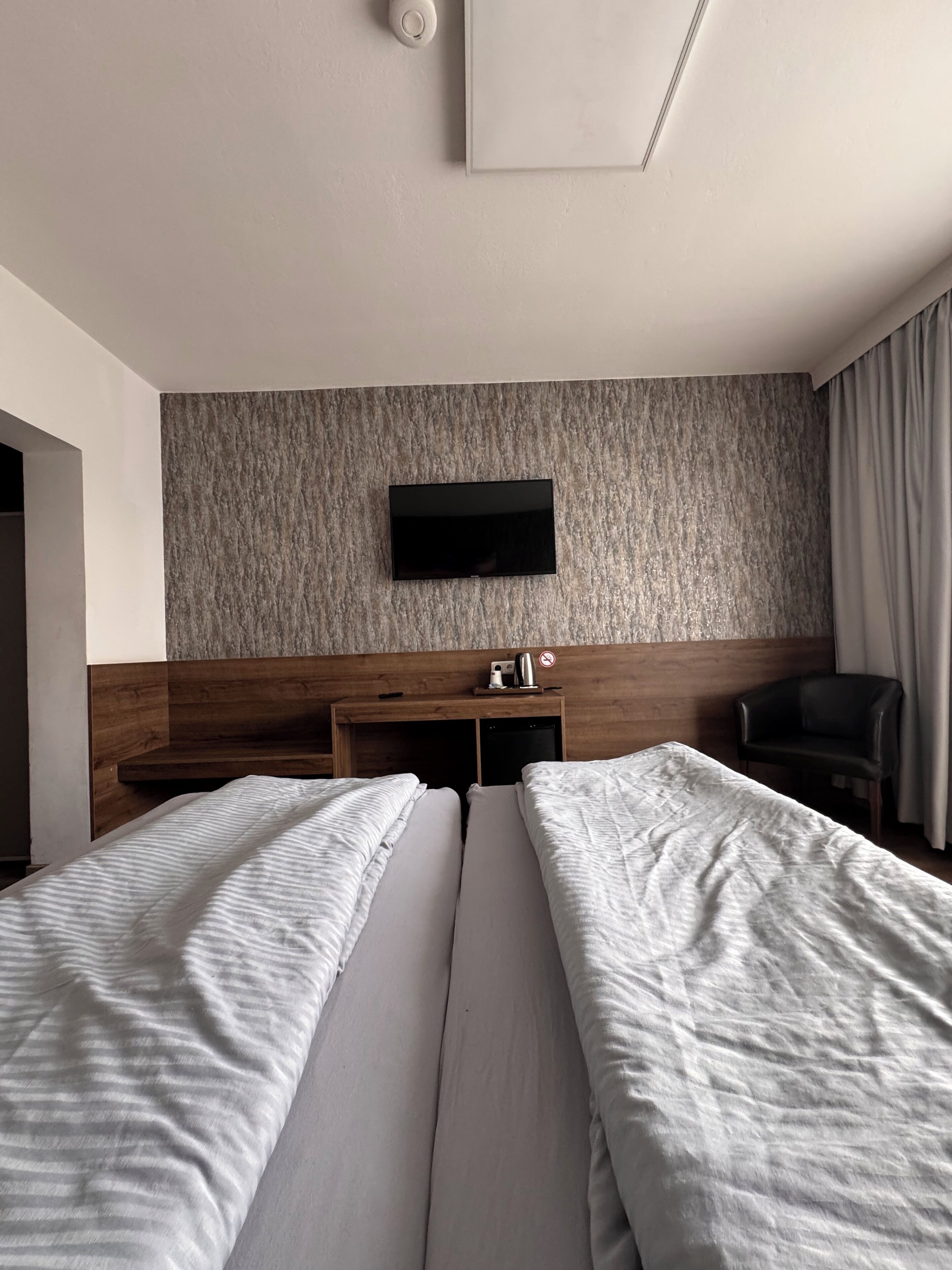 Une chambre d’hôtel avec deux lits, un téléviseur à écran plat fixé à un mur texturé, une tête de lit en bois, un bureau avec une bouilloire et un téléphone, et une chaise.