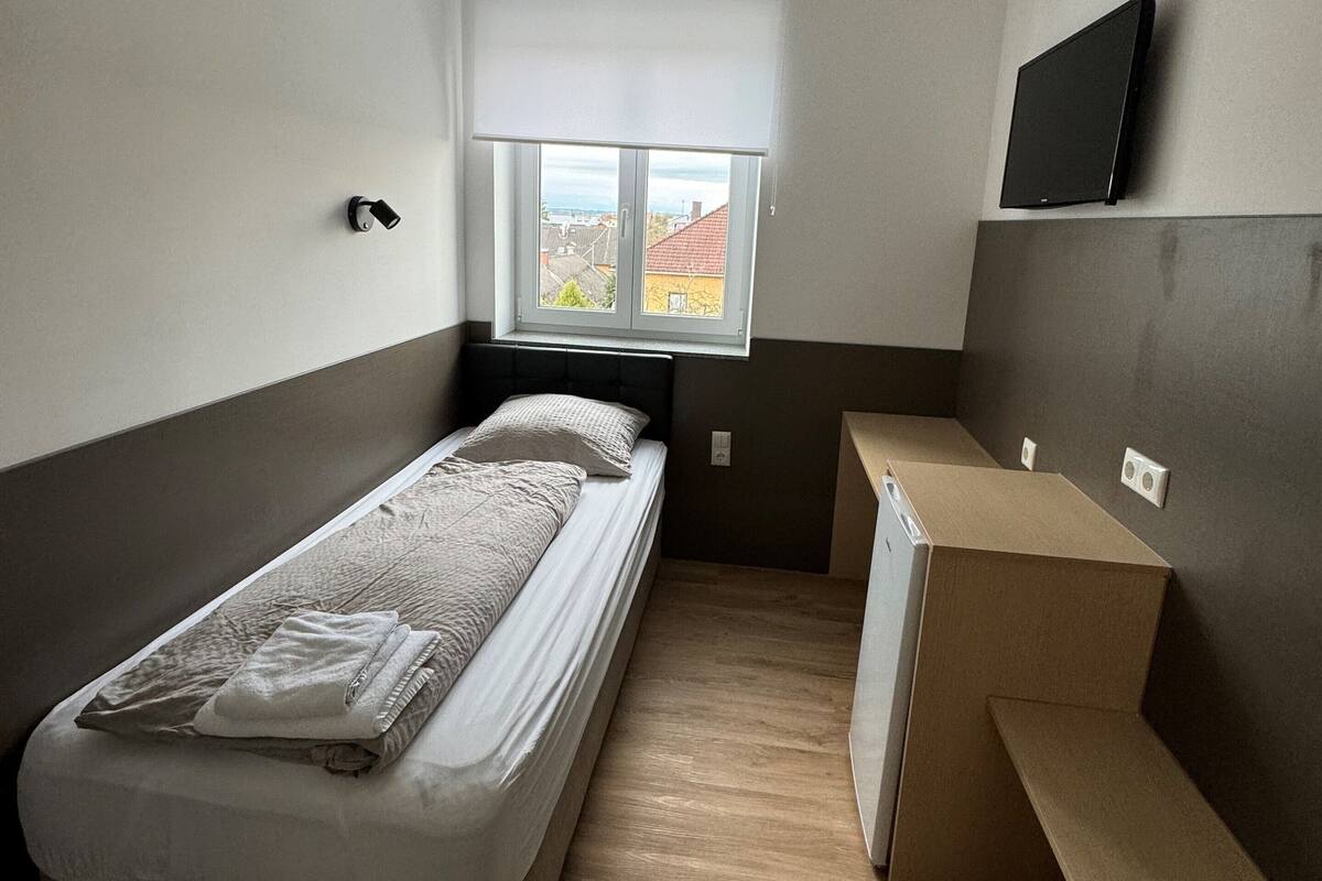 A compact room with a bed, a small desk, a window with a view, and a TV mounted on the wall.