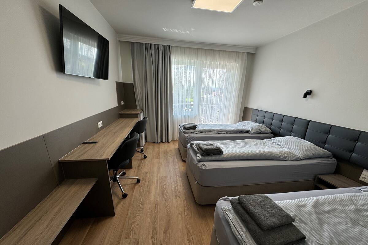 A hotel room with a wooden desk, two beds, a flat-screen TV, and a large window with curtains.