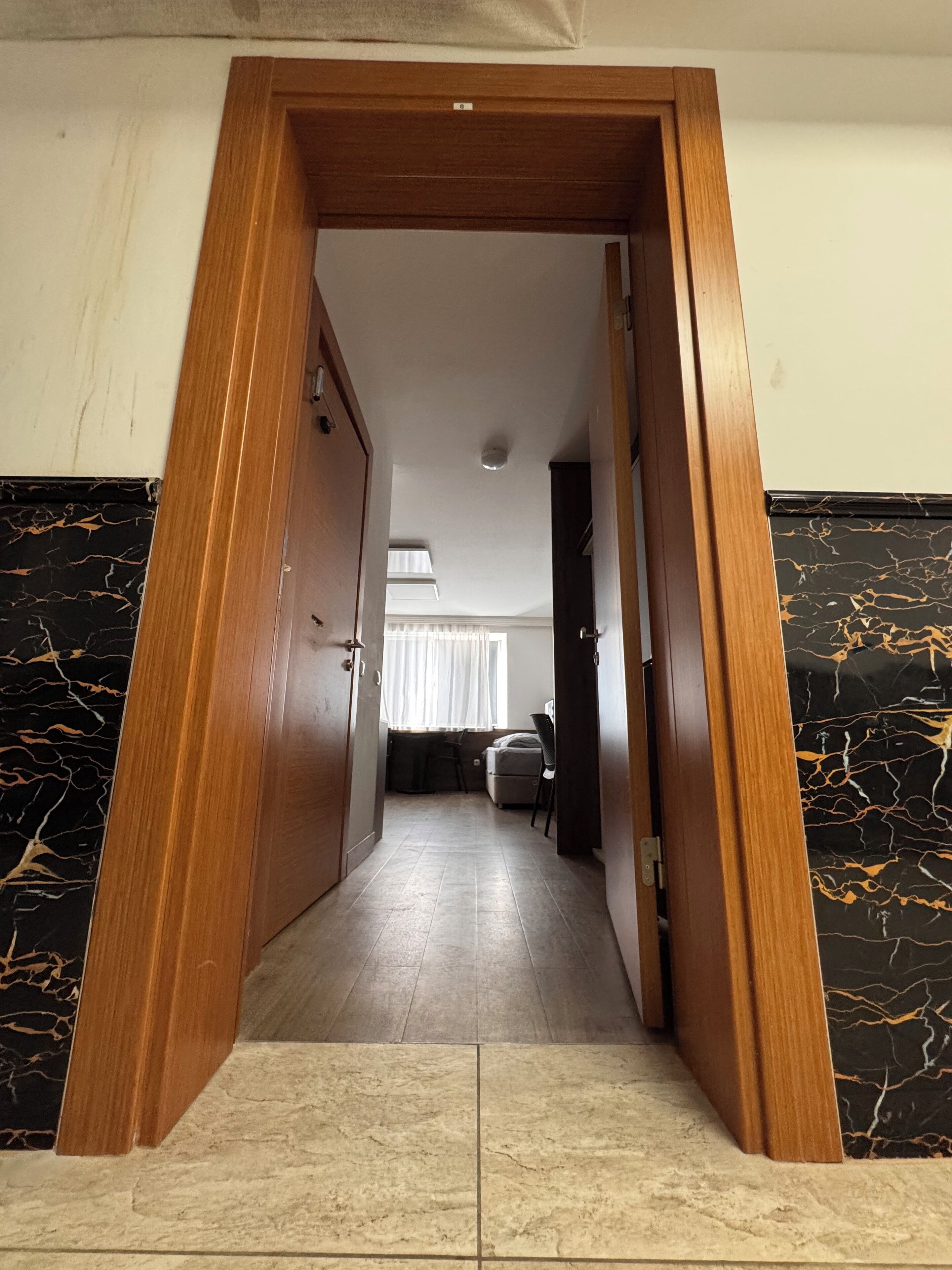 Un pasillo con marco de puerta de madera, suelo de baldosas y vista a una habitación con cama y sillón.