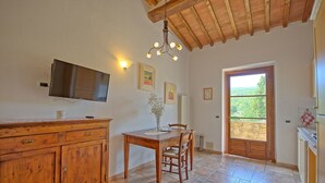 A kitchen with wooden cabinets, a dining table, chairs, a TV, and a window with a view of greenery.