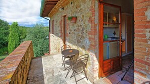 A stone building with a wooden door, a small patio with a table and chairs, and a view of greenery.