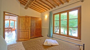 A bedroom with a large bed, a wooden wardrobe, a window with a view of greenery, and a door leading to a patio area. - Mulignone 1 - Apartment with pool (Molino D'elsa)