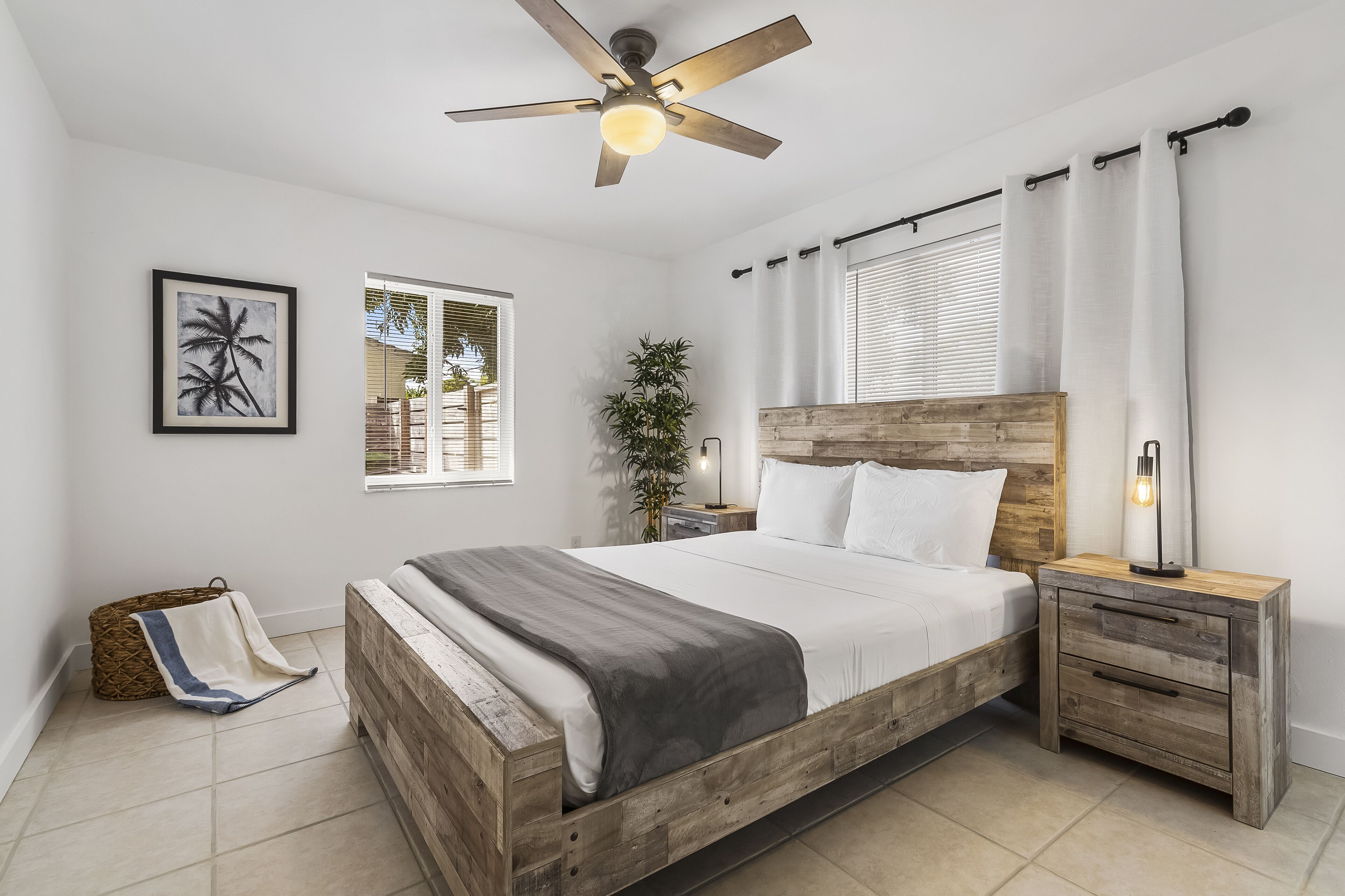 A bedroom with a wooden bed, a nightstand, a ceiling fan, a framed picture, a window, and a plant.