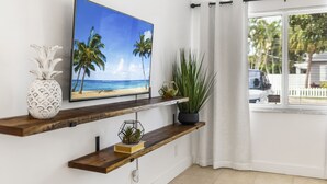 A room with a TV on a wooden shelf, a potted plant, and a view of the street through a window with curtains.