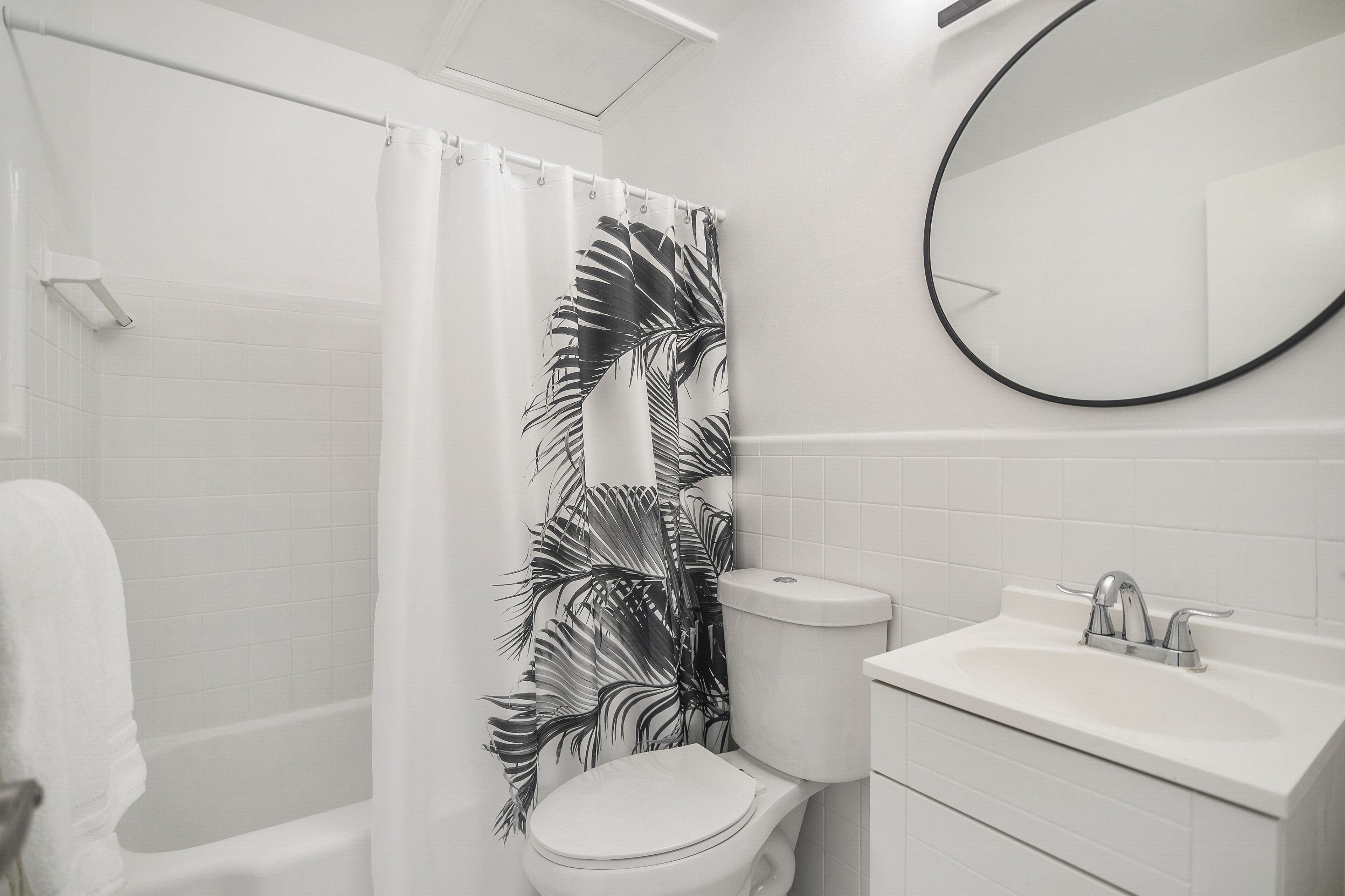 A bathroom with a shower curtain featuring a tropical leaf pattern, a toilet, a sink with a mirror above it, and a towel rack.
