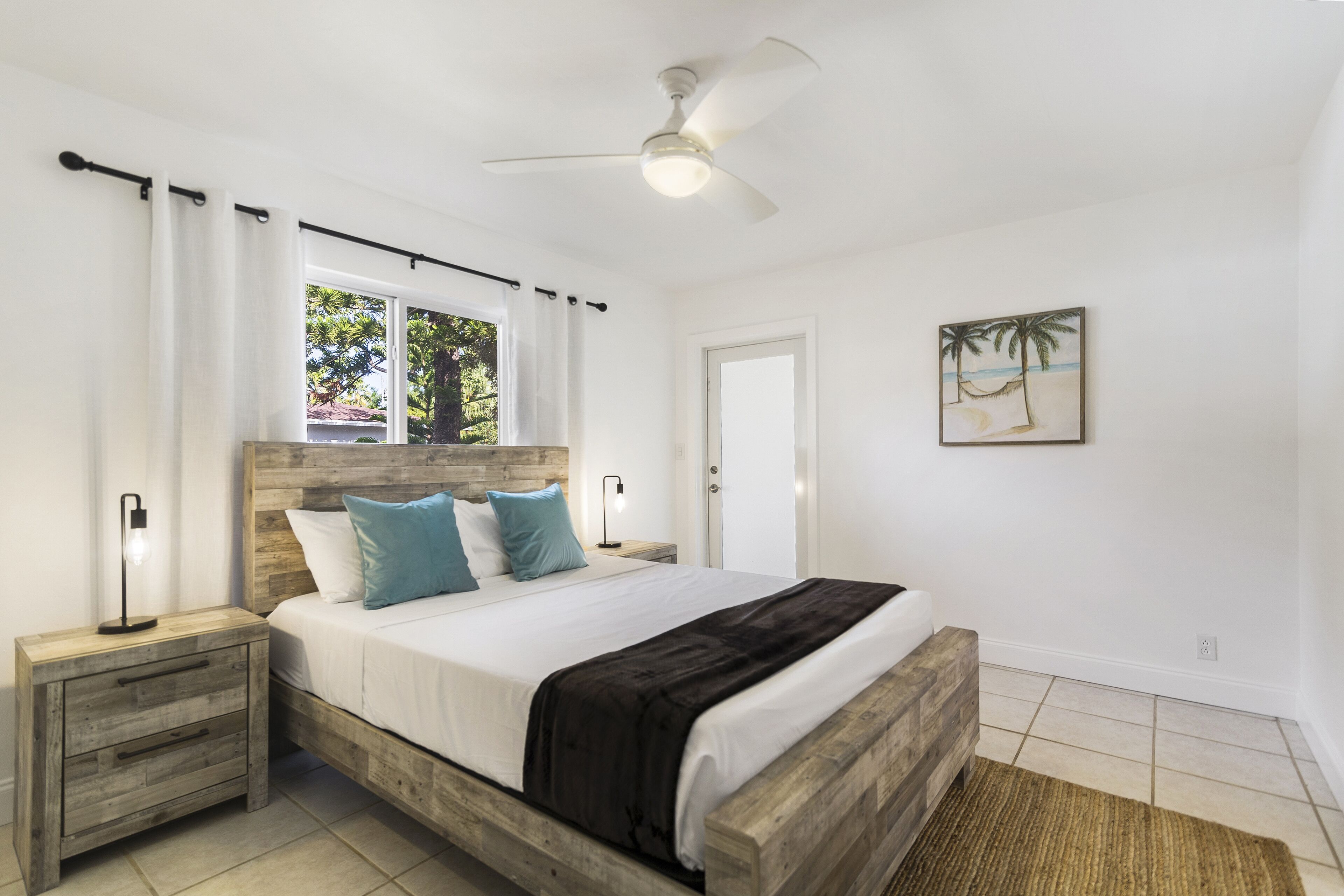 A bedroom with a wooden bed frame, white bedding, a ceiling fan, and a beach-themed painting on the wall.